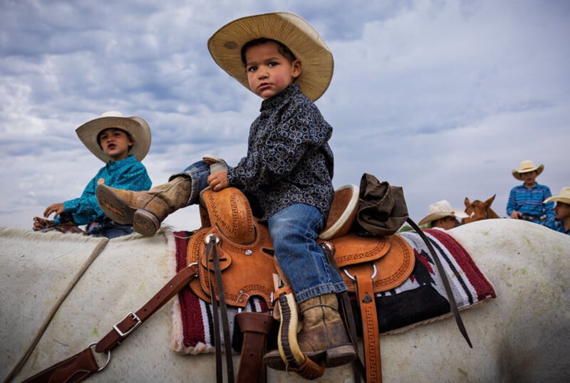 Un joven con sombrero y botas de vaquero se sienta en un caballo con arcos y mira a la cámara. Otro niño con traje y sombrero está cerca, con un cielo nublado de fondo.