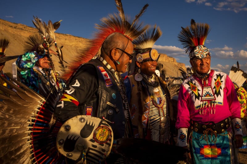 Cuatro hombres indígenas vestidos con coloridas ropas tradicionales y tocados de plumas están al aire libre, sonriendo y hablando, con la naturaleza, el paisaje soleado y el cielo azul de fondo.