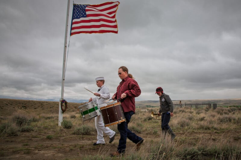 Tres hombres, entre ellos un marinero vestido de blanco y otros dos, caminaban por un campo de hierba llevando tambores y trompetas, con una enorme bandera estadounidense ondeando en un asta al fondo bajo un cielo nublado.