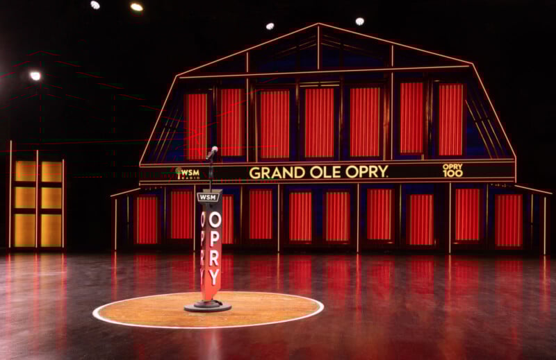 El escenario tiene un fondo de luz roja con forma de granero y un escenario marcado con un podio. "WSM Grand Olé Opry" y "opry 100," Hay un micrófono encima. El suelo es de madera pulida.