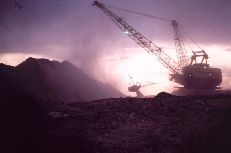 Una gran excavadora trabaja en un paisaje rocoso polvoriento al atardecer o al amanecer, recortada contra un cielo púrpura con montículos de tierra al fondo.