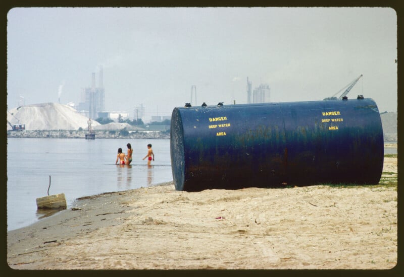 Un gran tanque de agua azul está etiquetado "Peligrosas aguas profundas" Sentados en la playa, tres niños chapotean en las aguas poco profundas cercanas; Al fondo se ven claramente edificios industriales y pilotes.
