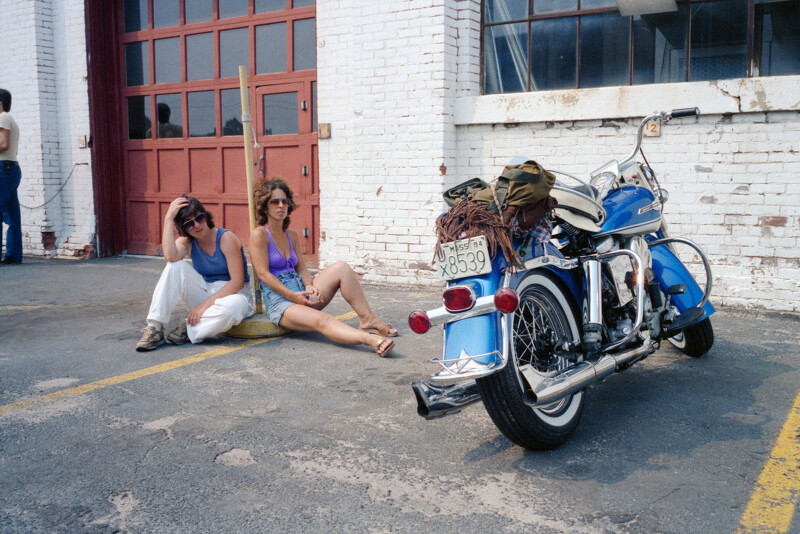 Two people sit on the ground outside a white brick building near a red garage door, with a blue motorcycle parked in front of them. The pair appears relaxed, wearing casual summer clothes.