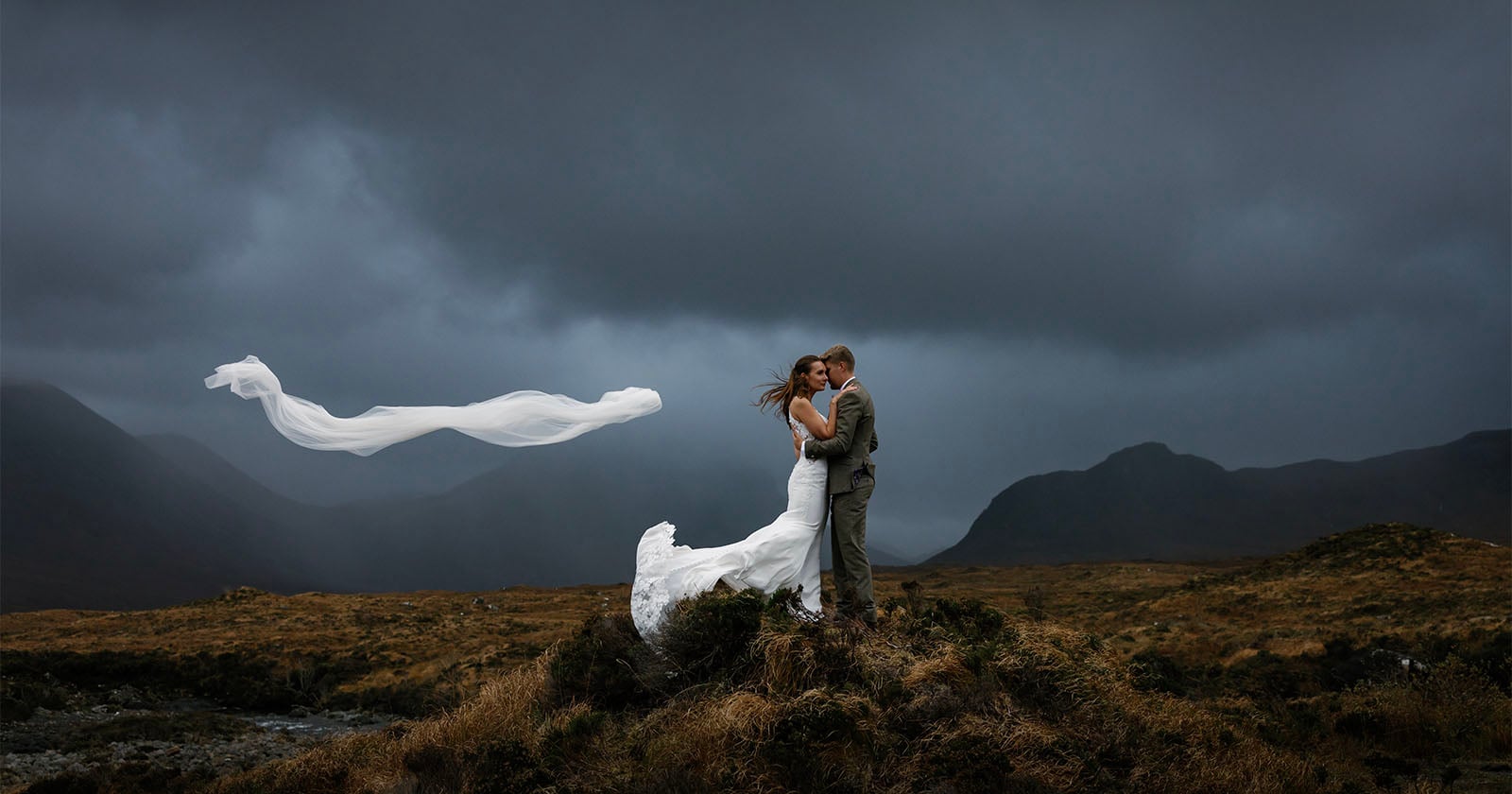 A bride and groom embrace on a grassy hilltop under a dramatic, cloudy sky. The bride’s veil is blowing in the wind, creating a flowing shape behind them. Rolling hills and mountains form the moody background.