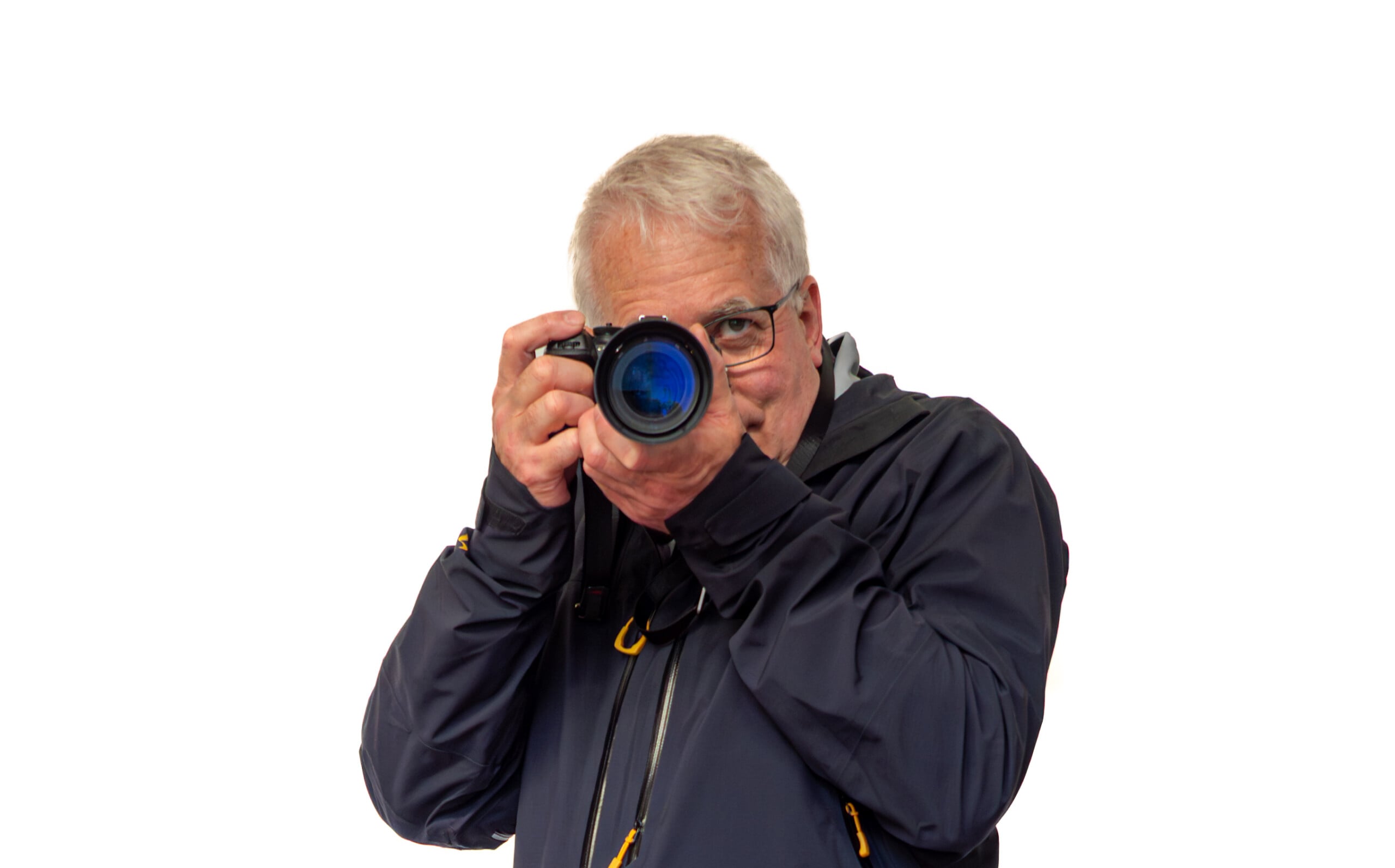 A man with gray hair and glasses, wearing a dark jacket, holds a camera up to his face as if taking a photo, against a plain white background.