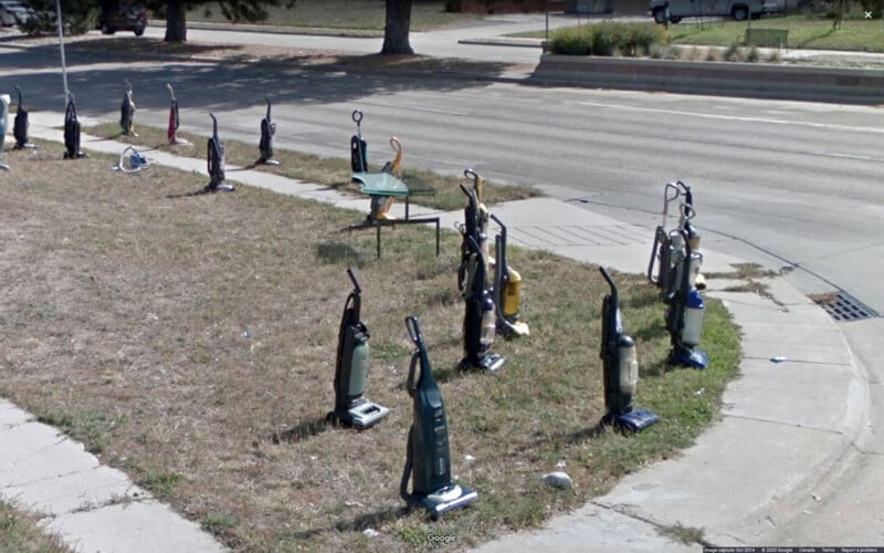 Decenas de aspiradoras verticales se encuentran en fila en el césped junto a la calle, con un pequeño cartel verde y adornos de flamencos en el fondo.