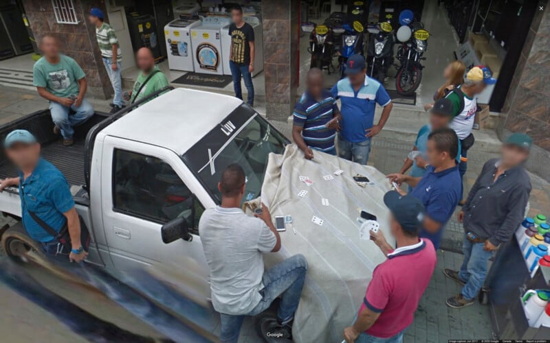 Un grupo de personas se reunió alrededor de una camioneta blanca estacionada en la calle, jugando a las cartas sobre una tela extendida sobre el capó. Al fondo se ve a varios curiosos y una motocicleta aparcada.