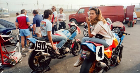 A woman sits on a colorful motorcycle, looking at the camera, while several people gather around her and another rider on a motorcycle marked "190" in a parking lot near a red van. Racing and casual atmosphere.