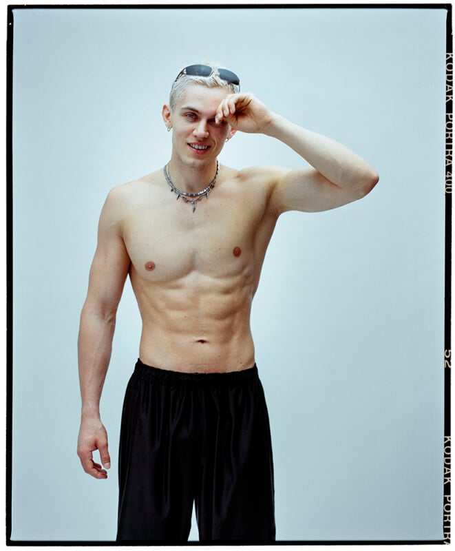 Un joven sin camisa y con el pelo claro se alza sobre un fondo claro, sonríe y se acaricia las cejas con una mano. Lleva un collar y gafas de sol en la cabeza.