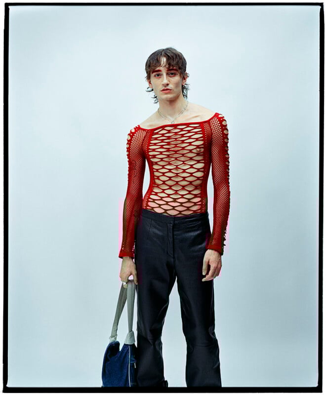 Un hombre con el pelo despeinado, vestido con una camiseta roja de manga larga con estampado de cruces, pantalones negros y un bolso gris azulado, estaba de pie contra un fondo claro y liso.