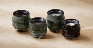 Four camera lenses on a wooden surface; three are green with white markings and one is black, all featuring various focal lengths and aperture values.