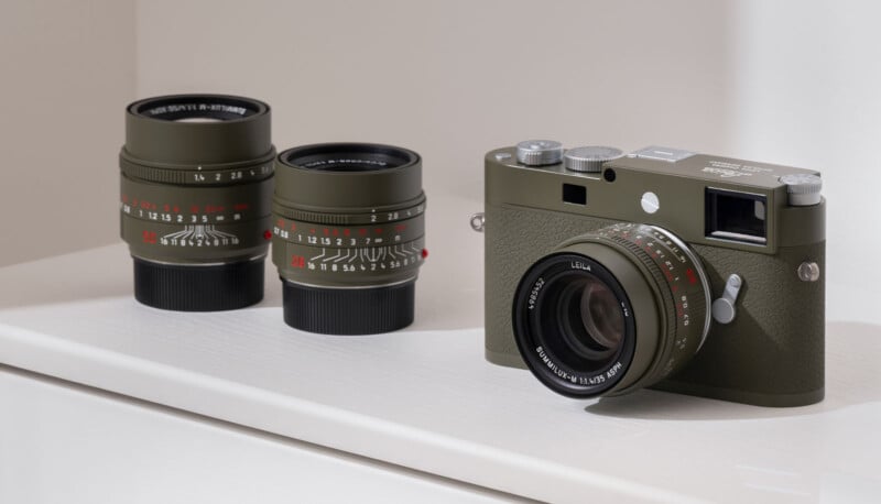 Se muestra una cámara Leica verde oliva con lente sobre una superficie blanca, junto a dos lentes de cámara intercambiables adicionales con apertura visible y marcas de enfoque.