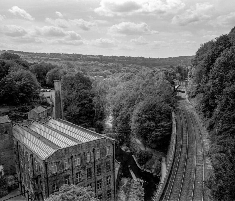 Black and white photo of an old industrial building near a railway track, surrounded by dense trees and rolling hills under a partly cloudy sky. The scene is peaceful and slightly elevated.