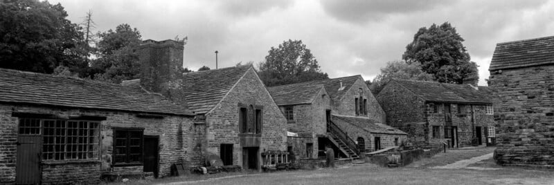 A row of old stone buildings with slate roofs on a grassy area, surrounded by trees under a cloudy sky. The scene appears quiet and historic.