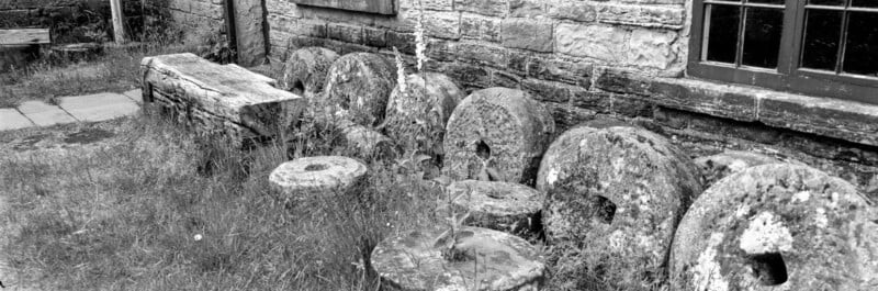 A row of old millstones leans against a rustic stone building with a wooden window. An aged wooden bench sits nearby, and grass grows among the stones, giving the scene an abandoned, historical feel.