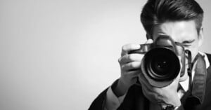 A man in a suit holds a DSLR camera up to his face, looking through the viewfinder as he takes a photo. The image is black and white, with a plain background.