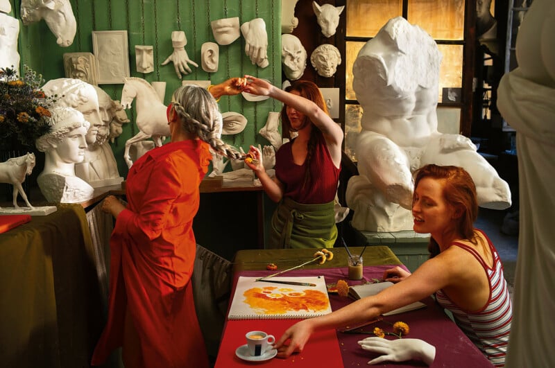 Three women are in an art studio filled with white plaster sculptures and busts. One woman braids another’s gray hair while the third, seated, sketches flowers, with art supplies and an espresso cup on the table.