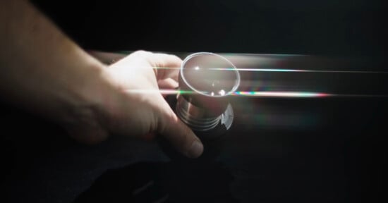 A hand holding a metallic cup against a dark background, with beams of light and subtle rainbow reflections passing through the scene.