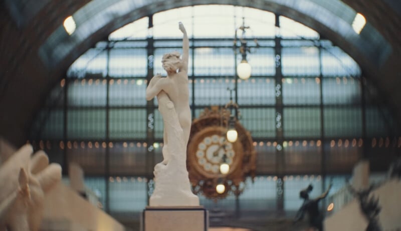 Una estatua de mármol blanco con los brazos levantados se encuentra en el gran salón, que tiene una gran ventana arqueada y un reloj ornamentado al fondo. La iluminación de la escena es suave y elegante.