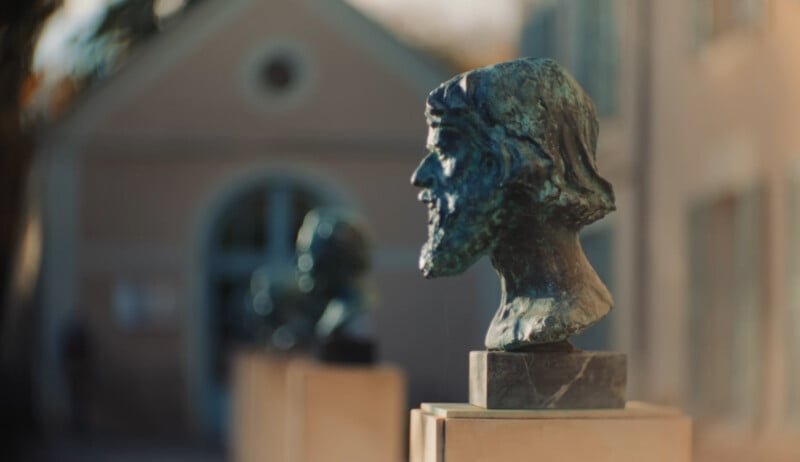 Un busto de bronce de un hombre barbudo se exhibe sobre un pedestal al aire libre, con otro busto y un edificio oscuro visible al fondo.