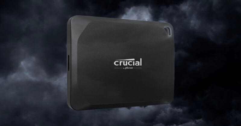 SSD externo Crucial negro con bordes redondeados mostrado frente a un fondo oscuro y nublado. El logotipo de Crucial aparece en fuente blanca en la parte frontal del dispositivo.