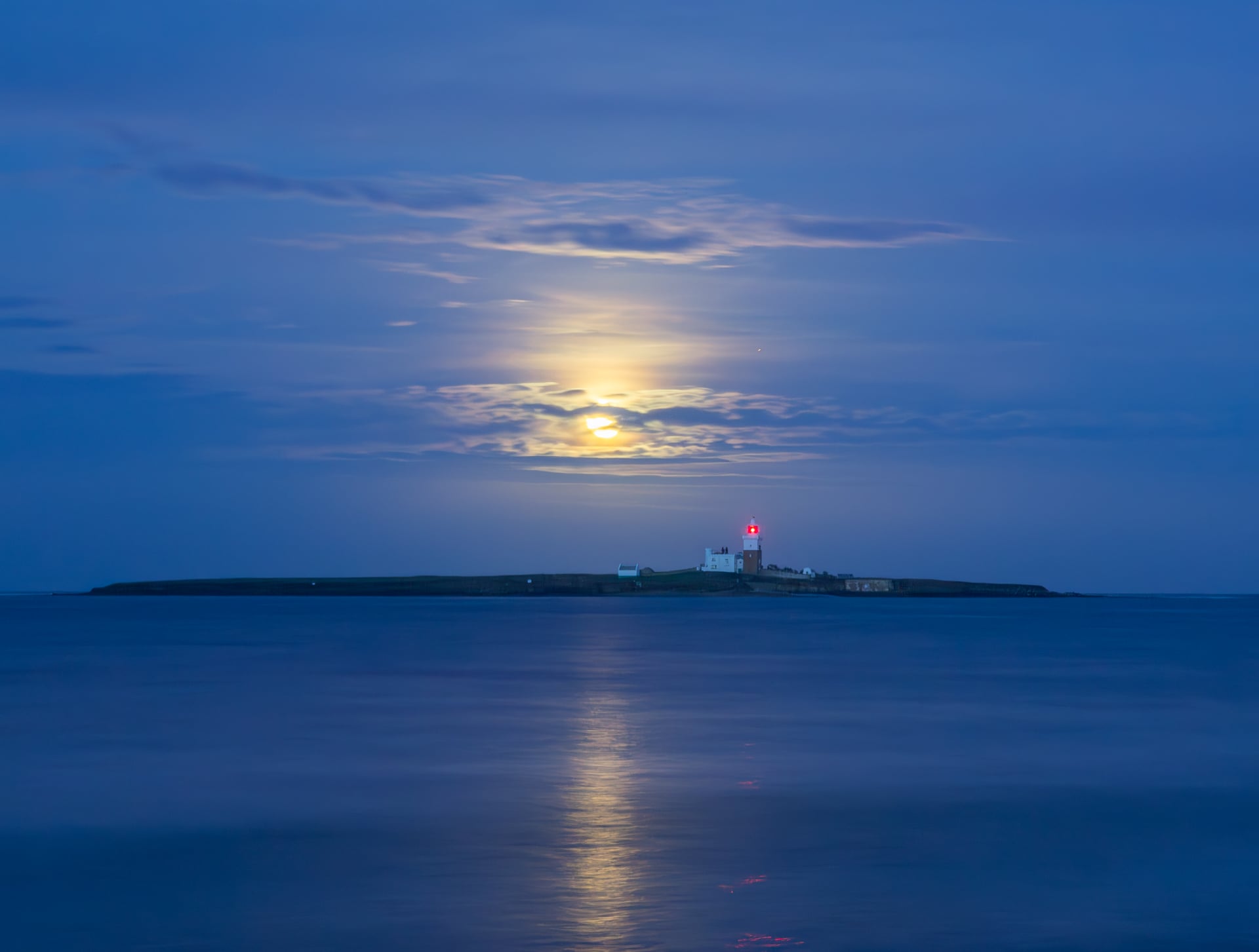 Bajo el cielo iluminado por la luna, una pequeña isla brilla con una luz roja y el reflejo del faro es claramente visible en el tranquilo mar azul.