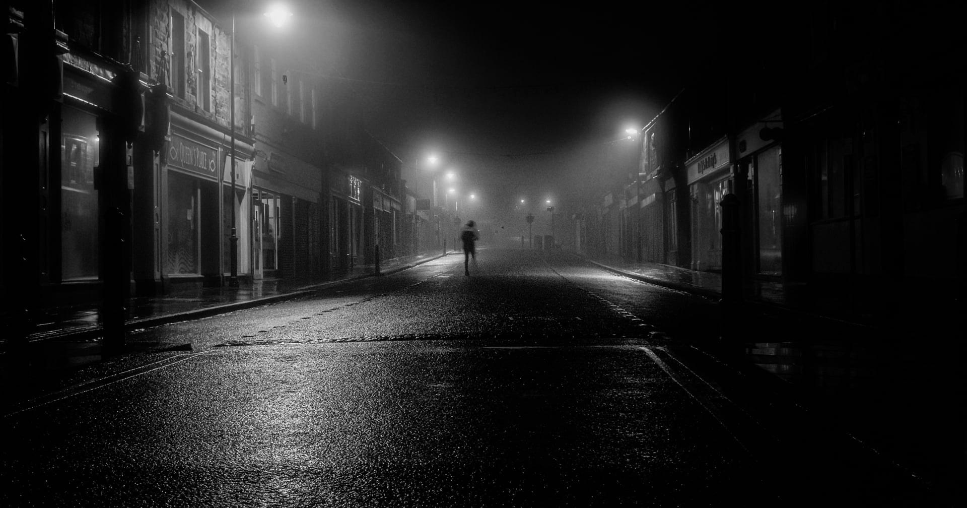 Por la noche, una figura solitaria camina por una calle vacía y húmeda, envuelta en niebla e iluminada por las farolas, creando una atmósfera misteriosa y de mal humor en un entorno urbano tranquilo.