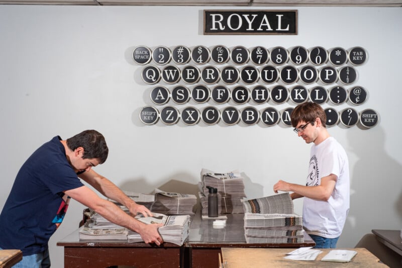 Two people sort newspapers on a table in front of a wall decorated with large round buttons arranged like a vintage typewriter keyboard. A "ROYAL" sign hangs above the buttons.