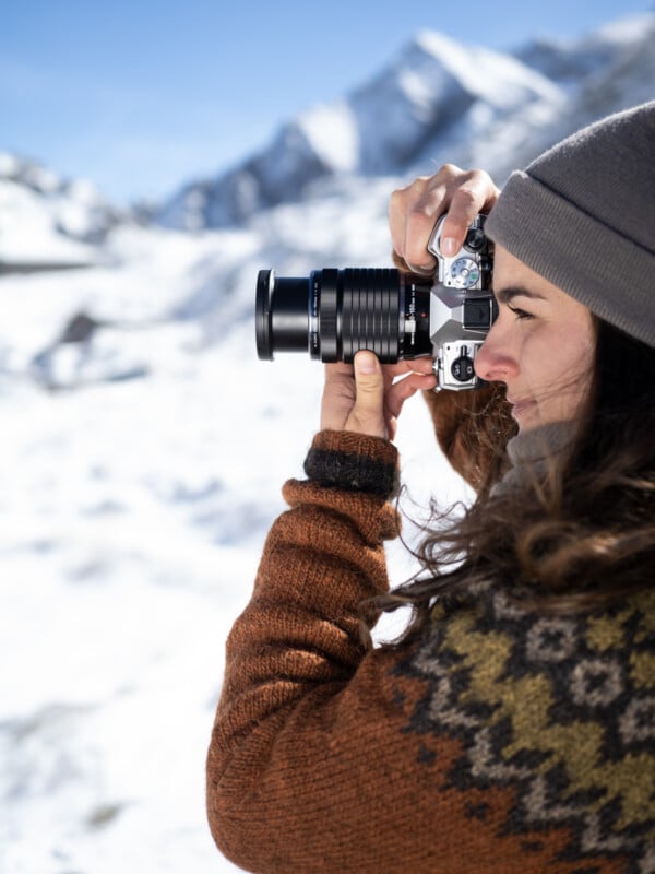 A person wearing a brown patterned sweater and gray beanie holds a camera, taking a photo in a snowy mountain landscape under a clear blue sky.