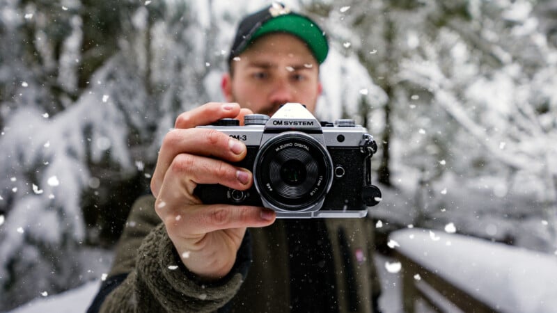 A person holding a silver and black camera toward the viewer, standing outdoors in a snowy forest. Snowflakes are falling, and the background is blurred with snow-covered trees.