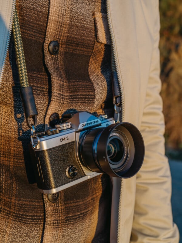 A close-up of a person wearing a plaid shirt and light jacket, with an OM System OM-3 camera hanging from their neck by a strap.