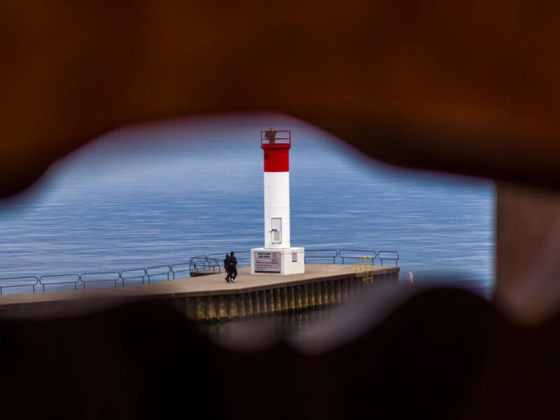 A person sitting near a red and white lighthouse at the end of a pier, seen through a blurred, irregularly shaped foreground frame, with calm blue water in the background.