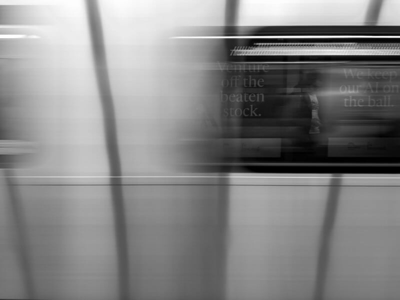 A blurred subway train passes by, with faint reflections of people and text visible through the window. Vertical lines from the platform or glass create a streaked effect, enhancing the sense of motion.