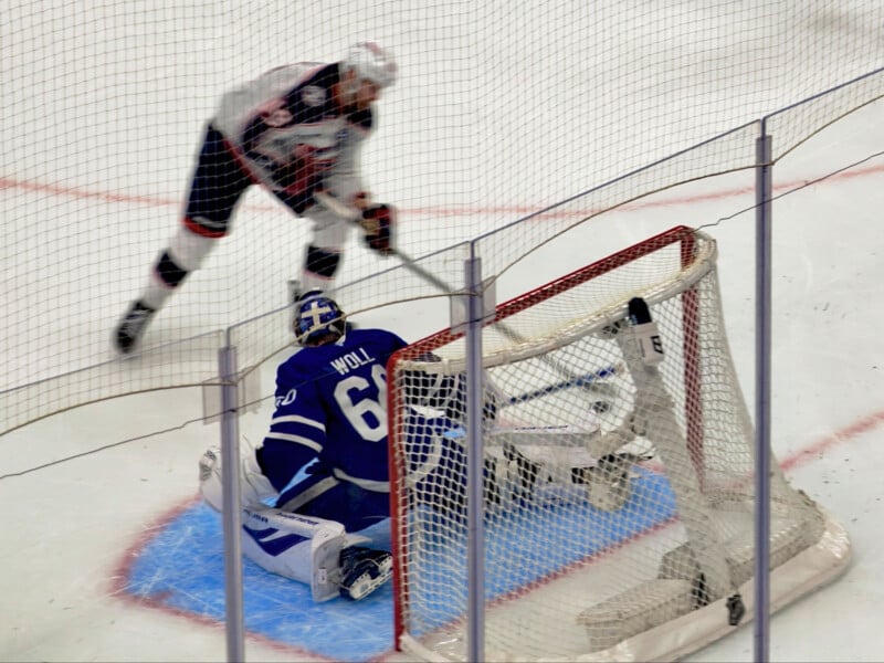 A hockey player in black and white gear takes a shot on goal while a goalie in blue and white gear, wearing number 60, attempts to make a save in front of the net.