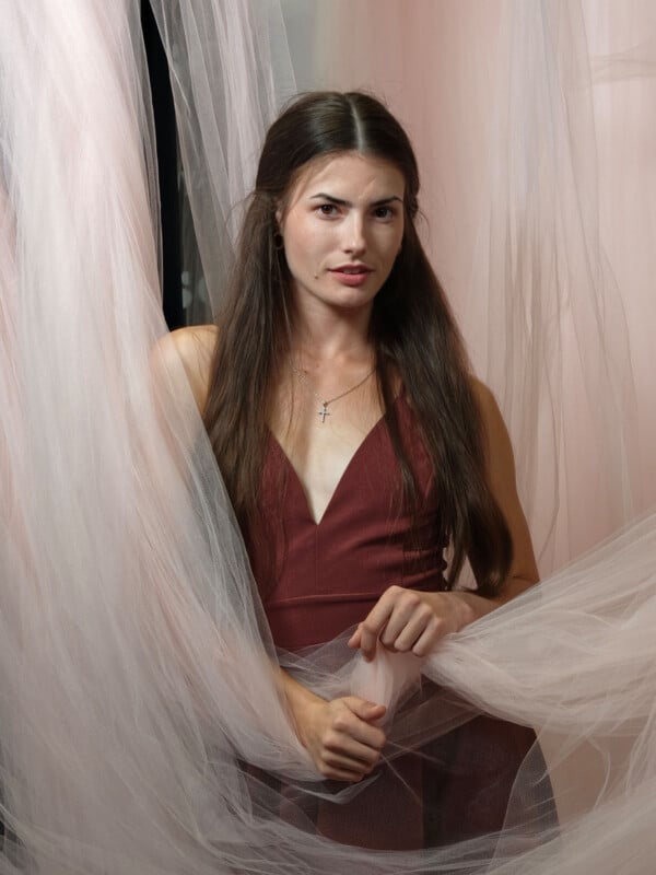 A woman with long brown hair wearing a sleeveless maroon dress stands among sheer pink fabric drapes, looking at the camera with a calm expression. She holds some of the fabric in her hands.