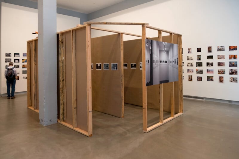 A wooden, unfinished room-like structure stands in a gallery. Photos are displayed on its walls and on the surrounding gallery walls. A person with a backpack views the photos on the far left wall.