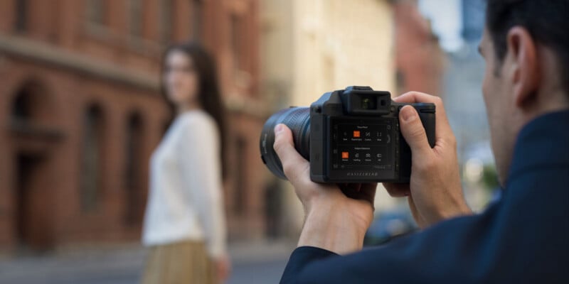 A photographer holds a digital camera and focuses on a woman standing outdoors. The woman in the background is blurred, while the camera’s screen and controls are clearly visible.
