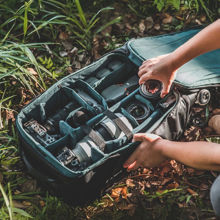 Una persona organiza el equipo fotográfico y las lentes en una bolsa acolchada para cámara mientras está al aire libre, rodeada de hierba y follaje.