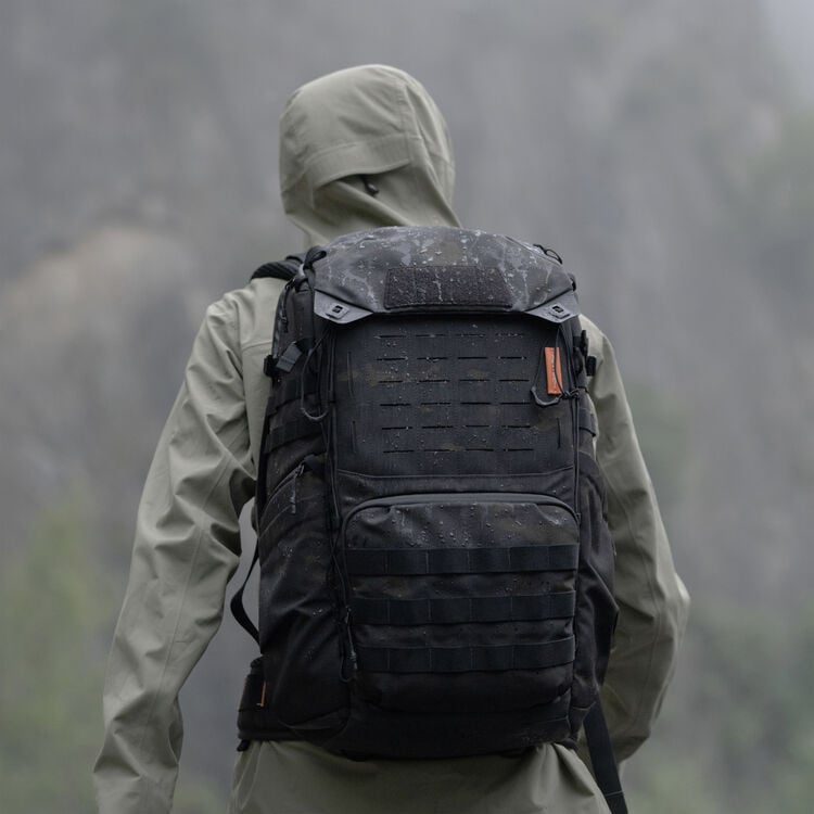 A person in a hooded, gray rain jacket stands outdoors with a large black backpack. The backpack is wet with raindrops, and the background is a misty, blurred natural landscape.
