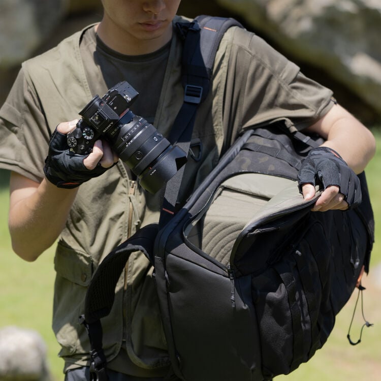 A person wearing fingerless gloves holds a camera in one hand and opens a black camera backpack with the other, standing outdoors on a sunny day.