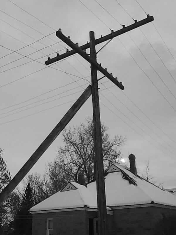 Una fotografía en blanco y negro de un poste telefónico con cables frente a una casa con el techo cubierto de nieve. Al fondo aparecen ramas desnudas y un cielo nublado.