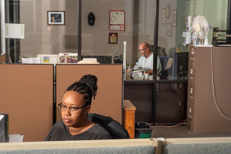 A woman with glasses works at a desk in an office cubicle, while a man sits at a desk in a separate office behind a glass partition filled with papers and folders. Both appear focused on their tasks.