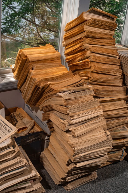Tall, uneven stacks of old, yellowed newspapers are piled by a window, with sunlight streaming through. The newspapers appear worn and fragile, and the surrounding area looks cluttered.