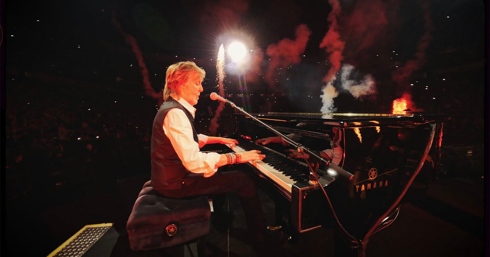 A musician plays a grand piano on stage, singing into a microphone. Fireworks and smoke light up the dark concert venue, and the audience is visible in the background. The piano brand "Yamaha" is seen on the instrument.