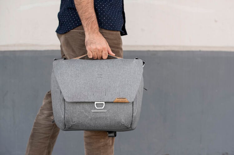 Un hombre vestido con pantalones marrones y una camisa azul marino se encuentra frente a una pared de dos tonos gris y blanco, sosteniendo una bolsa de mensajero gris por el asa superior.