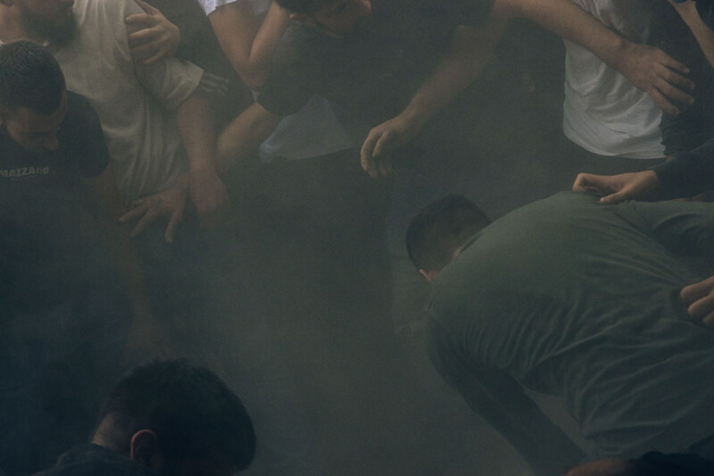 A group of people stand closely together in a smoky or dusty environment, some reaching out with their hands, their faces partly obscured by the haze.