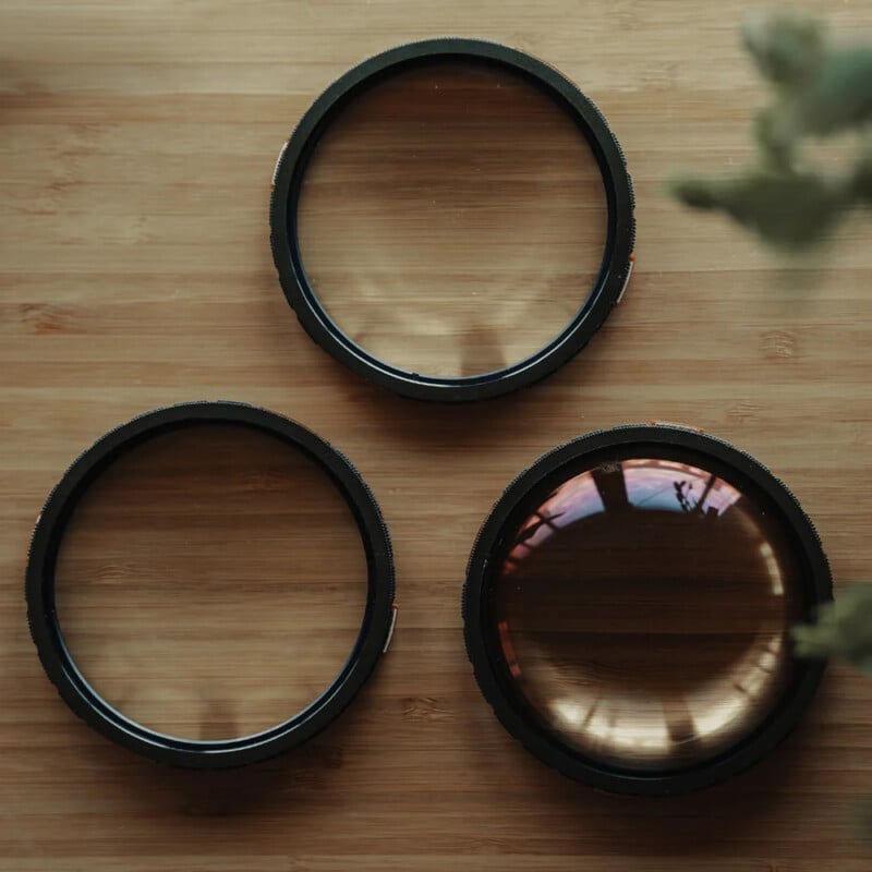 Three circular camera lens filters are arranged on a wooden surface, with two at the bottom and one at the top. Soft natural light and a blurred green plant are visible in the upper right corner.