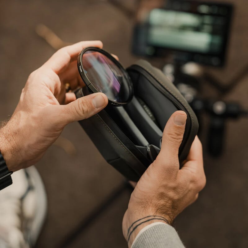 A person holds a zippered case containing camera lens filters, removing one circular filter. A camera and monitor are visible in the blurred background, suggesting a photography or videography setup.