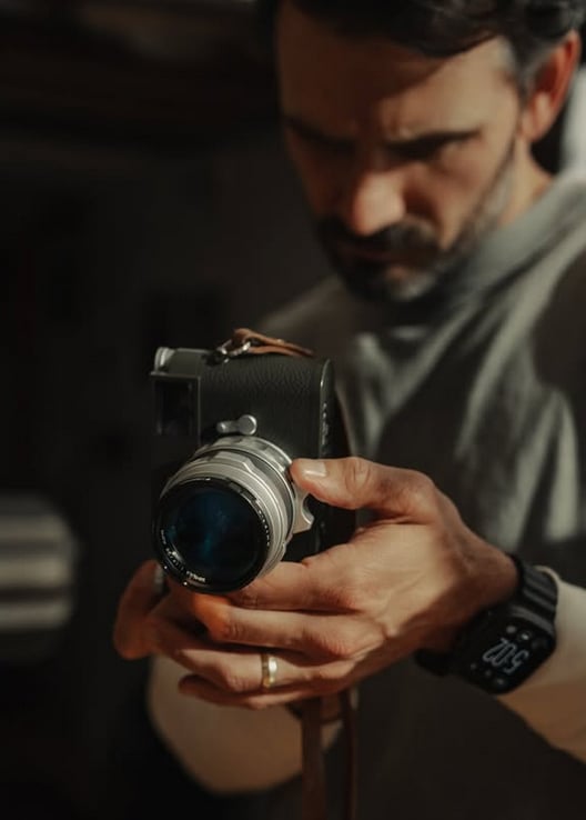 A man wearing a smartwatch holds a camera with both hands, focusing intently on the device. The background is softly blurred, drawing attention to the camera and his hands.