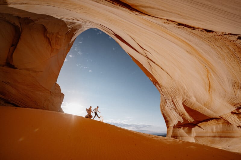 Dos personas se toman de la mano mientras caminan por una pendiente arenosa, que contiene un gran arco de roca iluminado por el sol con curvas y texturas dramáticas contra el cielo azul.